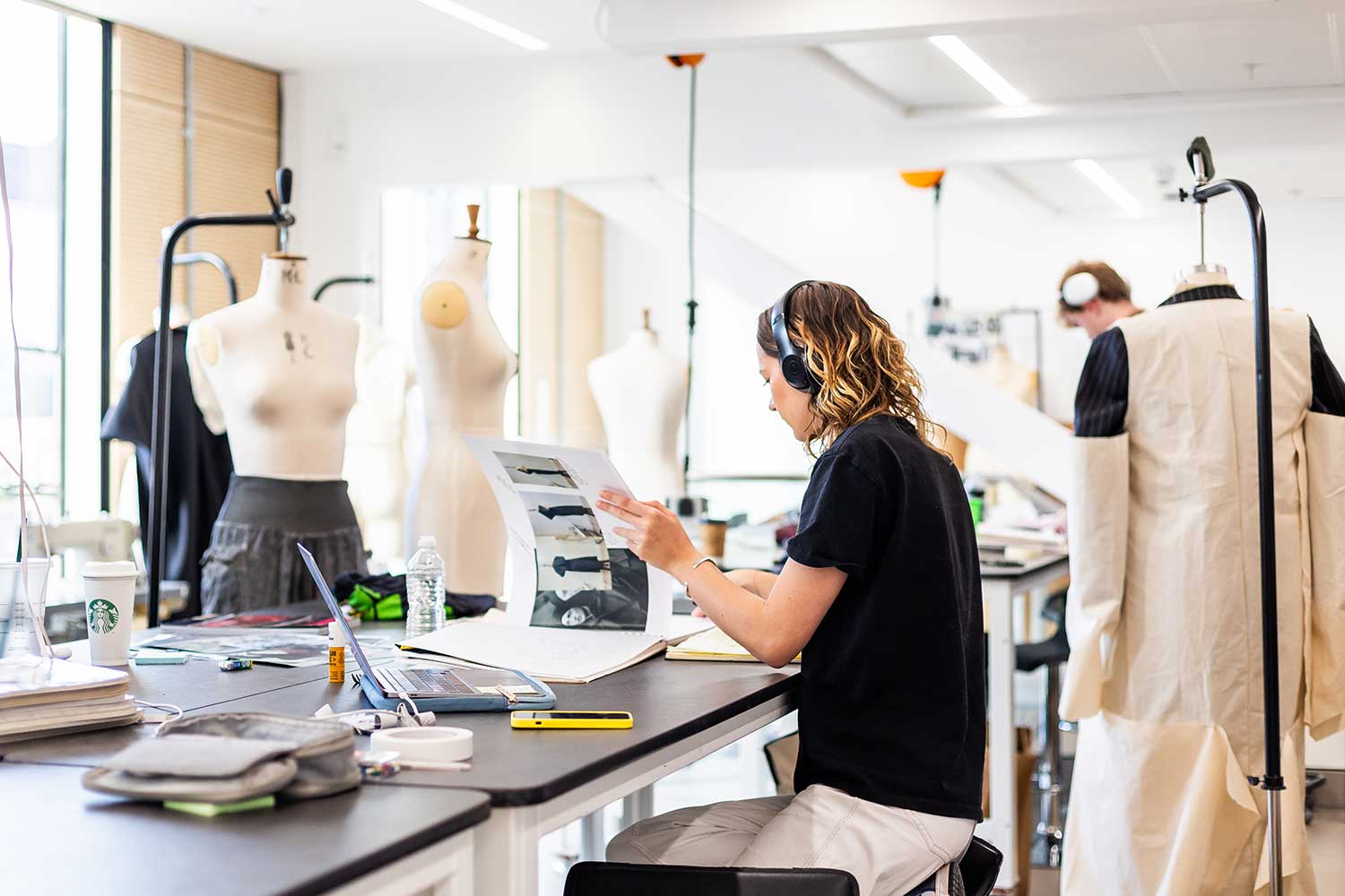 A student wearing headphones flips through a sketchbook while surrounded by mannequins