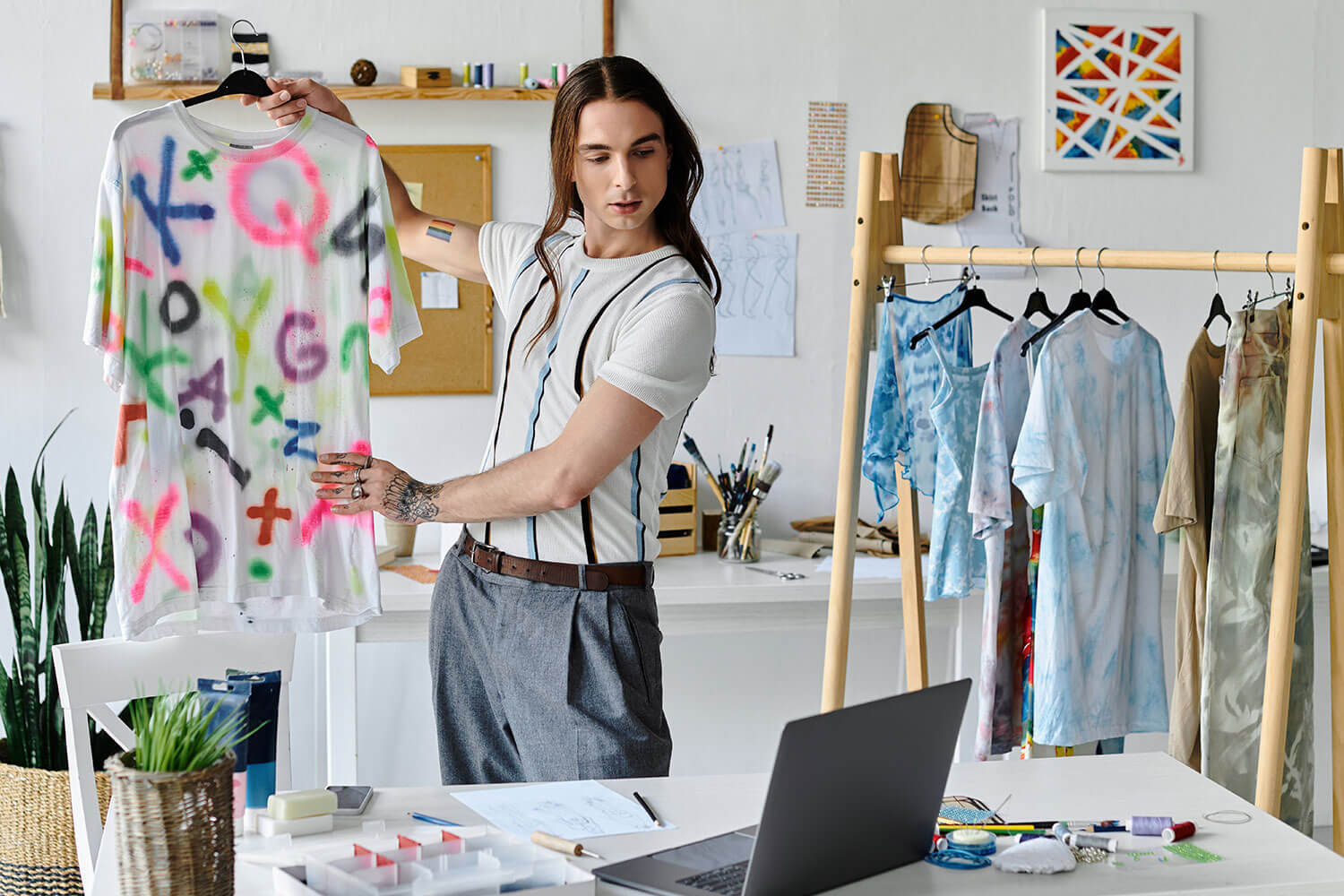 Person with long hair in a design studio holding clothes and looking at a laptop