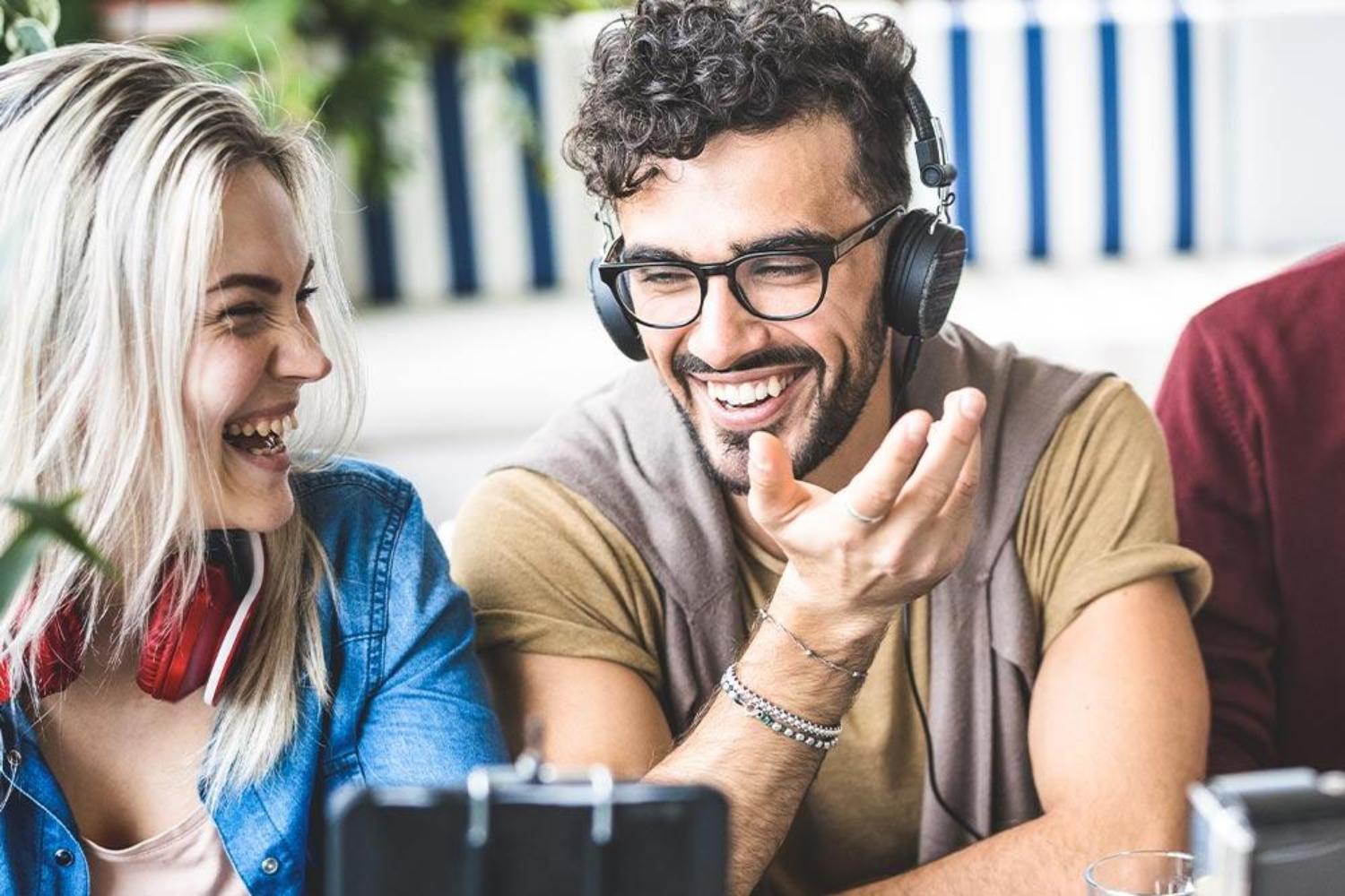 tWO YOUNG PEOPLE TALKING OVER COFFEE . mALE WEARING HEADPHONES AND THERE ARE CAMERAS ON STANDS IN FOREGROUND