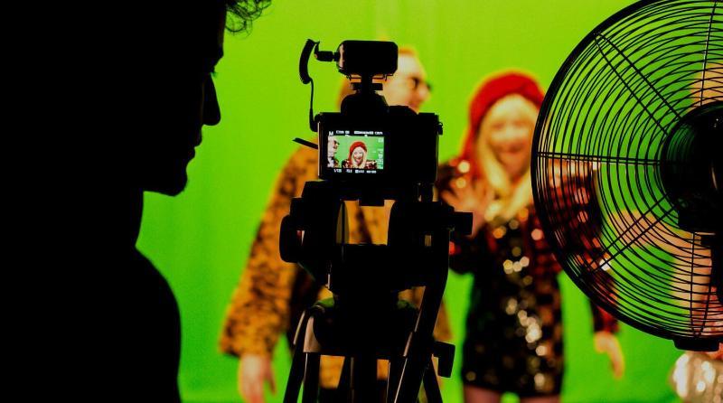 Silhouette of a student an camera on a tripod filming other students against a green screen. The students being filmed are out of focus but they are smiling and one is wearing a sequin dress and bright beret while the other is wearing sunglasses and a faux fur coat.
