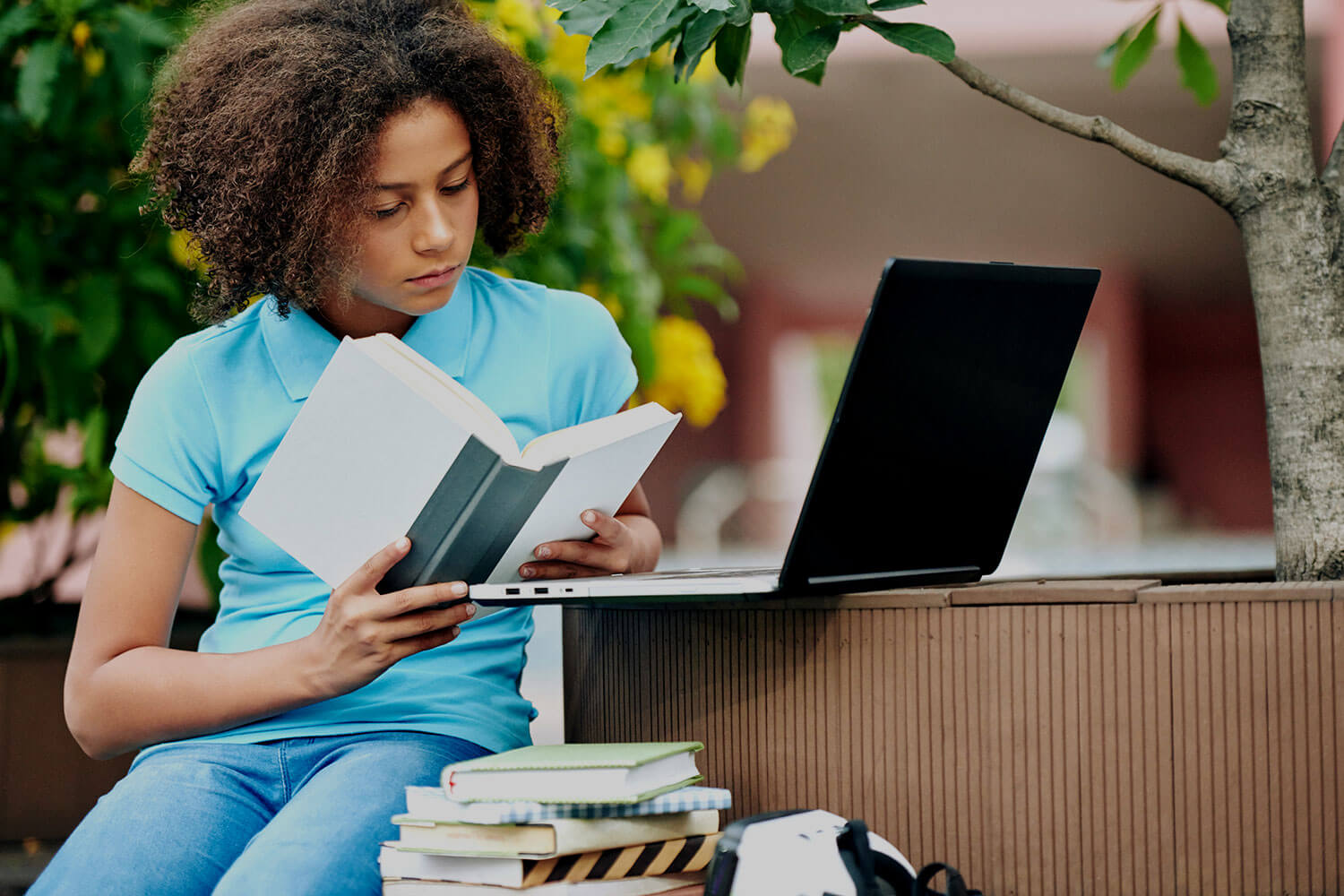 A girl sits outside reading a book with a laptop placed in front of her