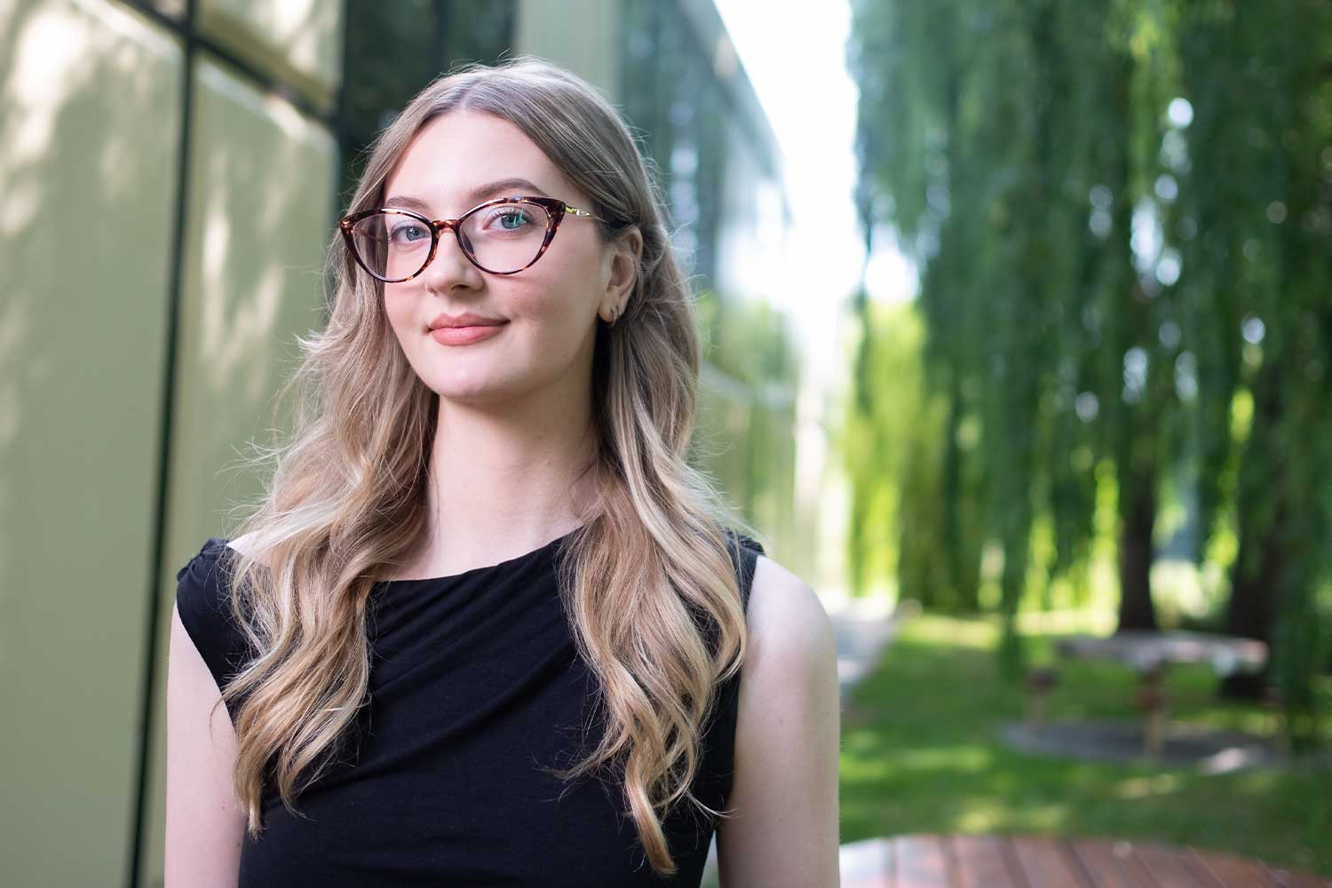 A person with long blonde hair and dark glasses stands outside a building surrounded by willow trees