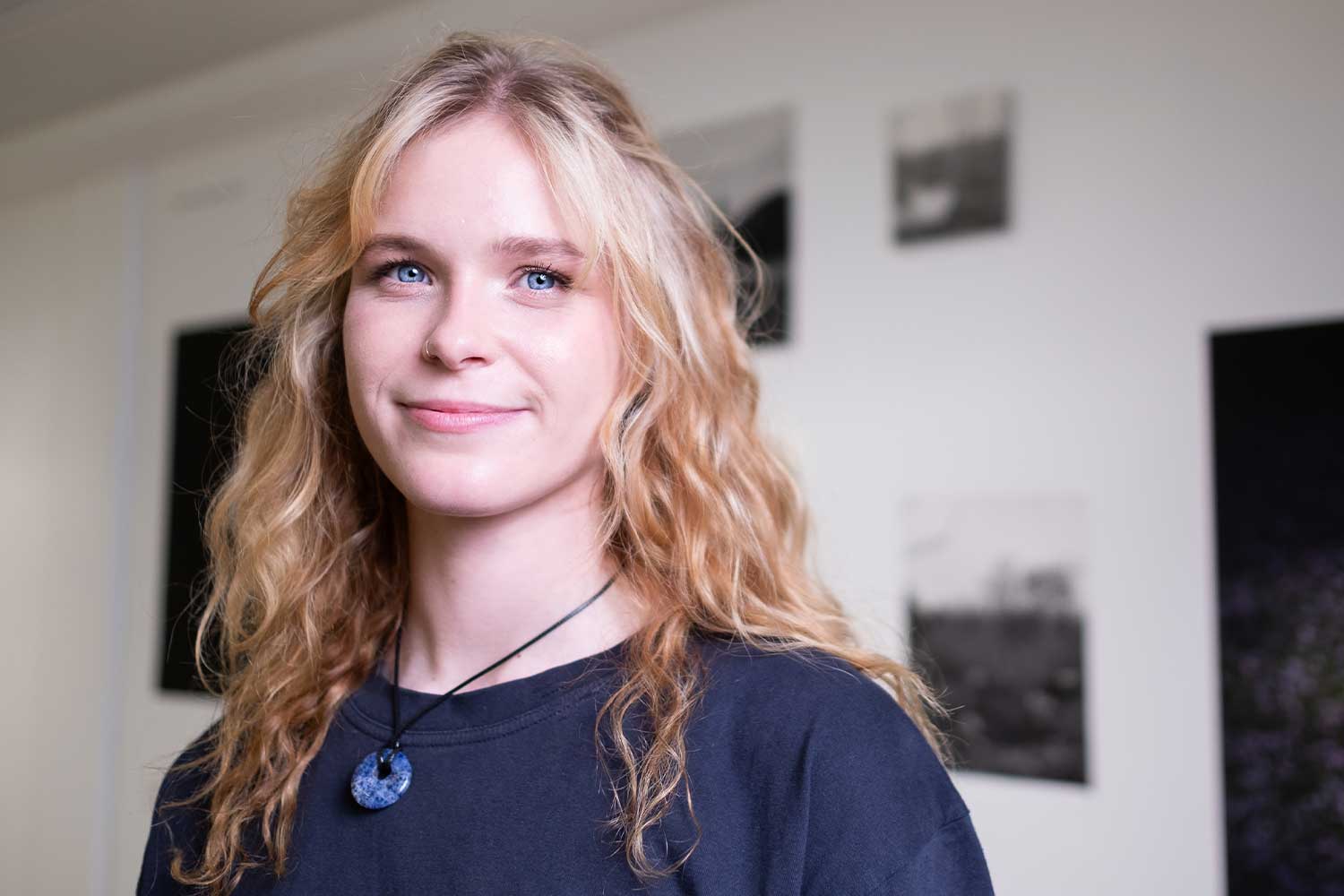 A person with long blonde curly hair and blue eyes smiles at the camera