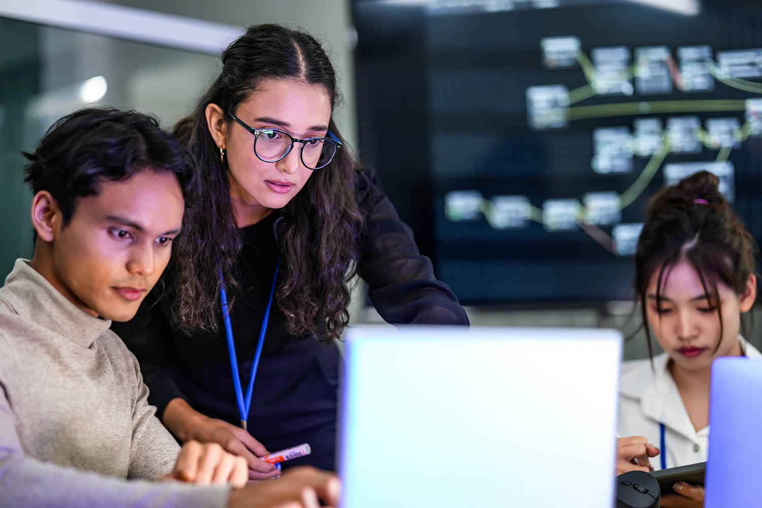 Three young people gathered around a laptop. Strings of coding on a screen in the background