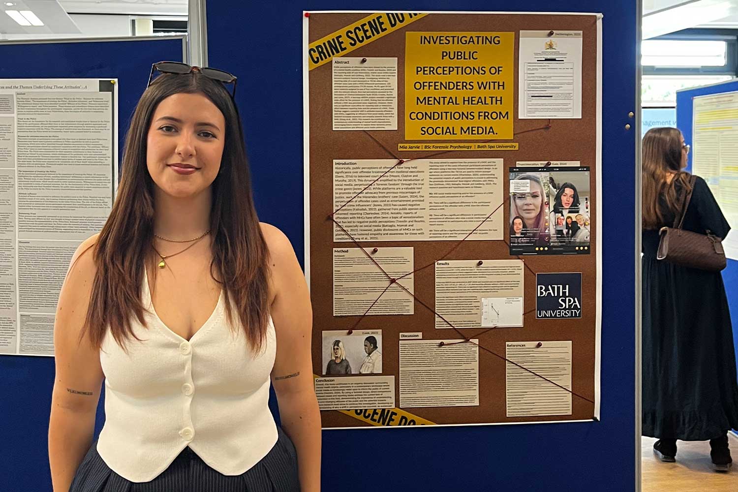 A student wearing a white waistcoat stands in front of a noticeboard showcasing their work