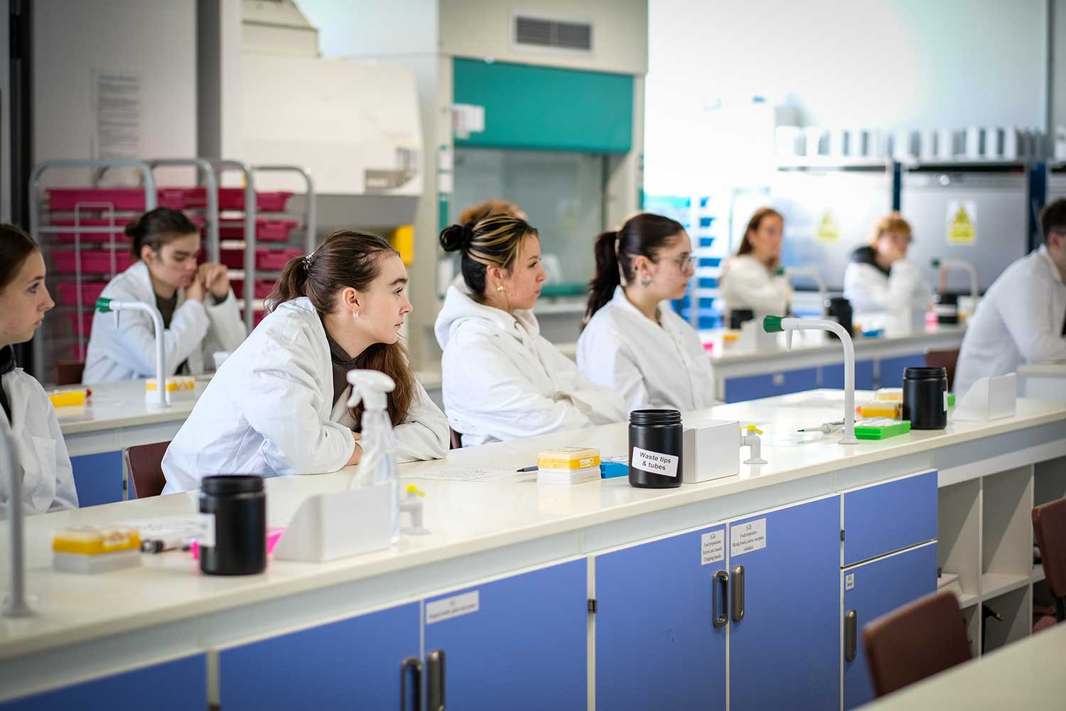Students wearing white lab coats listen to a lecturer speaking