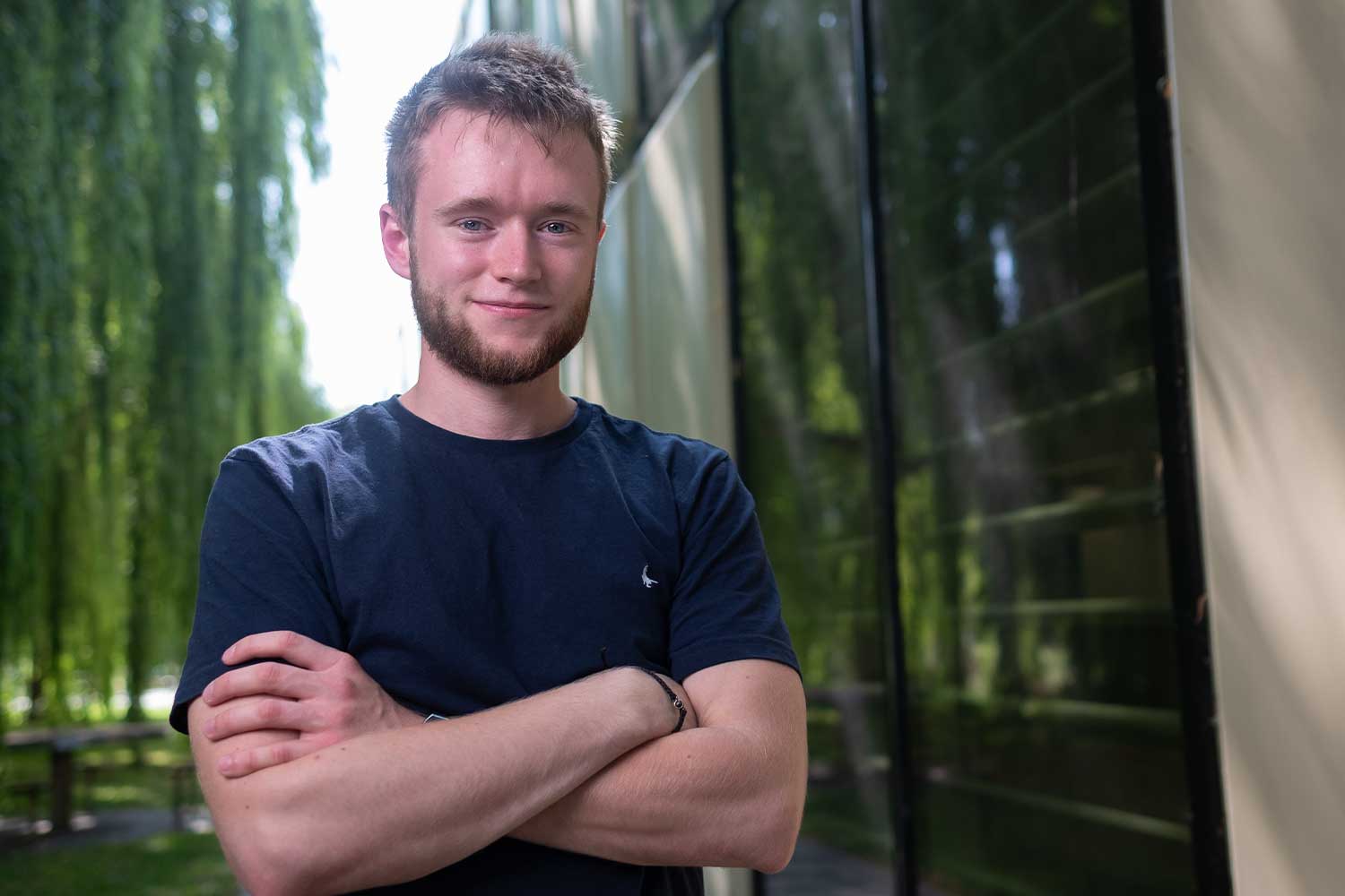 A person stands with their arms crossed in front a building surrounded by willow trees