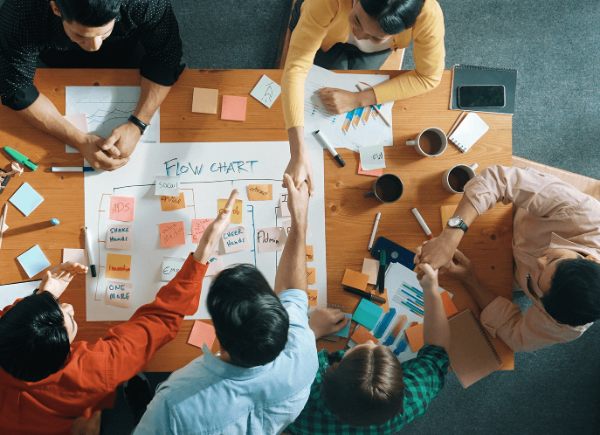 Six coworkers sit around a table shaking hands and working on a flow chart
