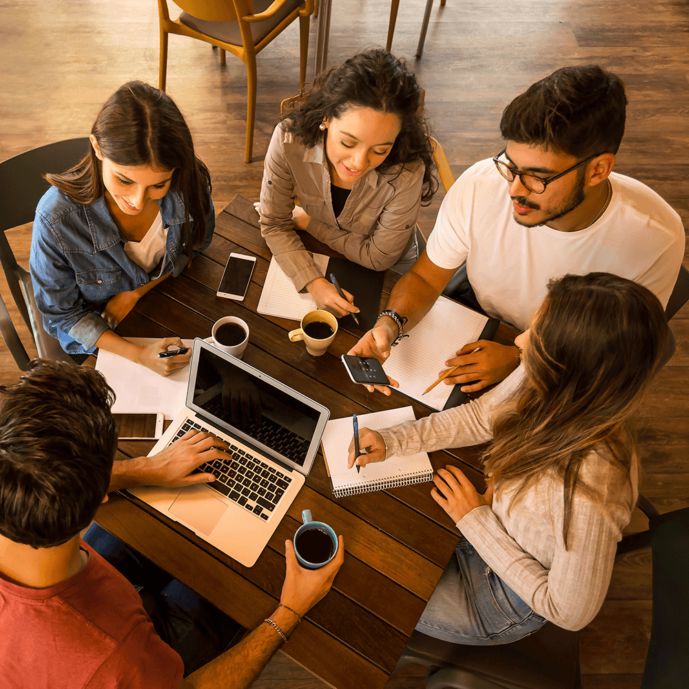Five students sit around a table drinking coffee and writing in notebooks