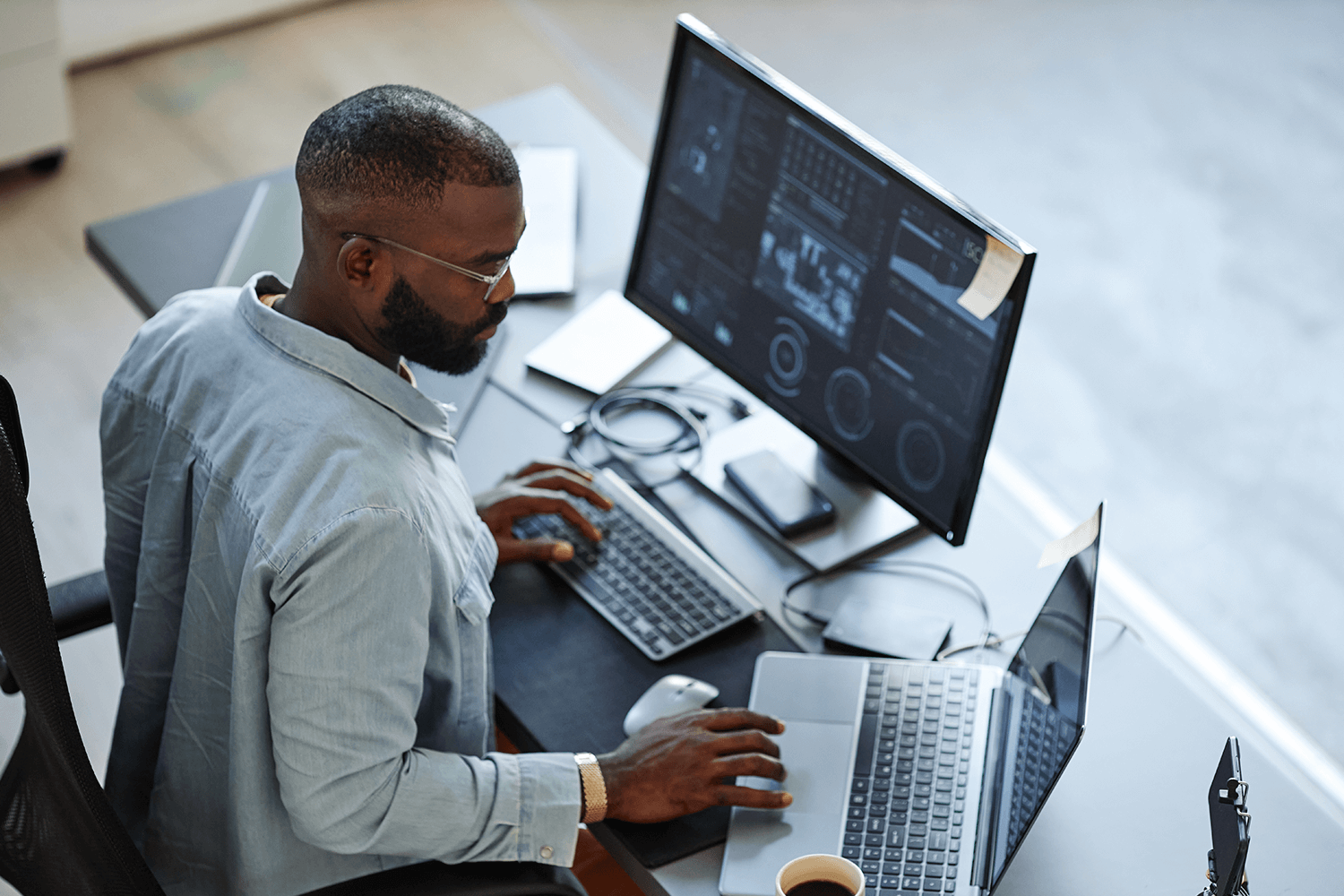A man sits at his desk looking at data dashboards on his laptop and monitor screens