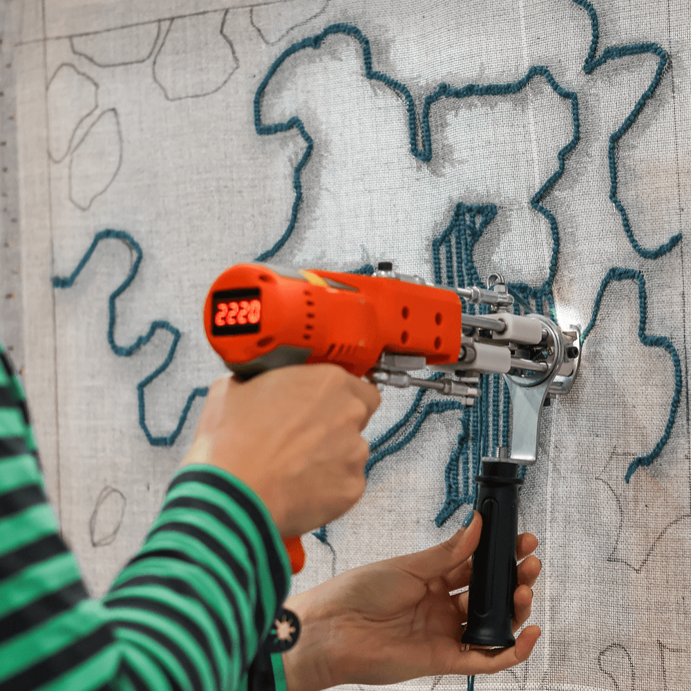 A student using an electric tufting gun to create the outline of a design in blue yarn on a piece of fabric.