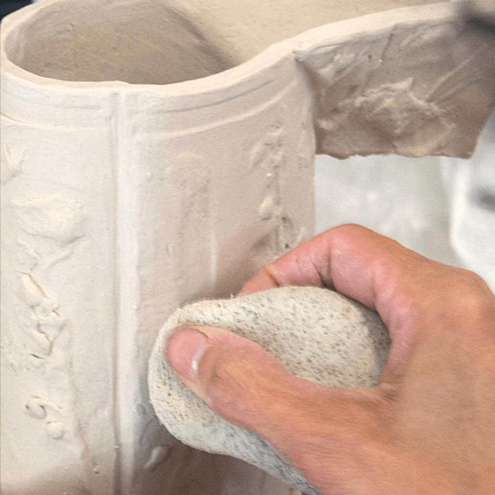 A hand uses a damp sponge to smooth out a ceramic jug