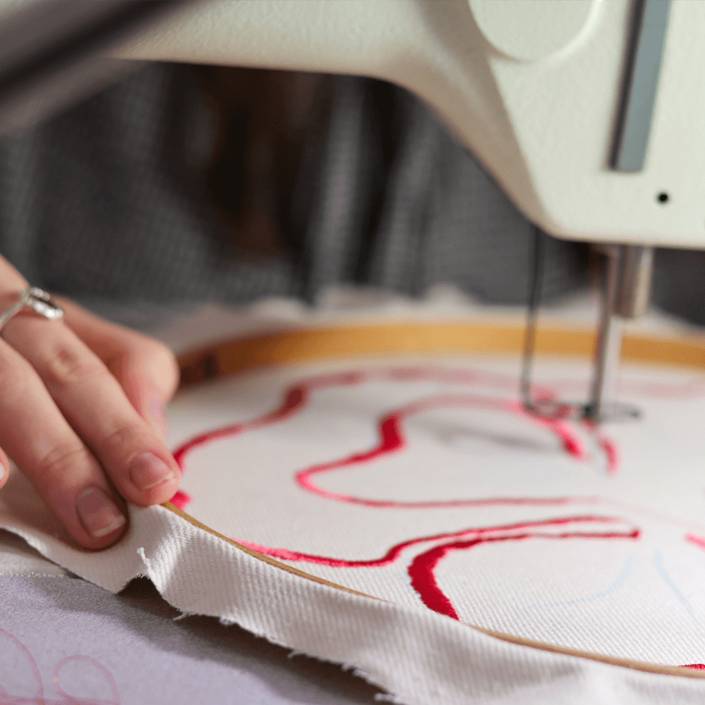 A student using a sewing machine to embroider a red freehand design onto a piece of cloth within a hoop