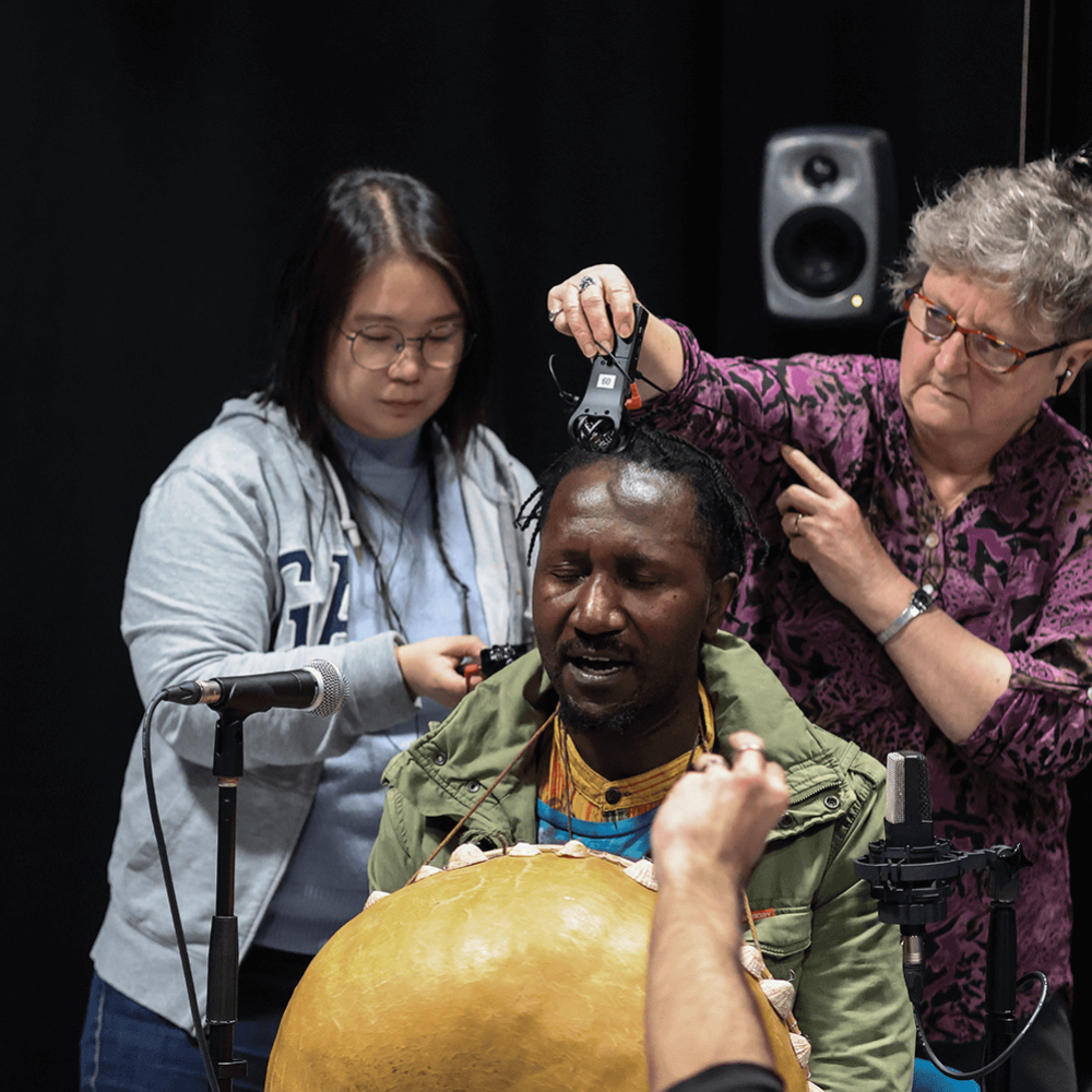 Students stand with sound recorders around a man playing an instrument