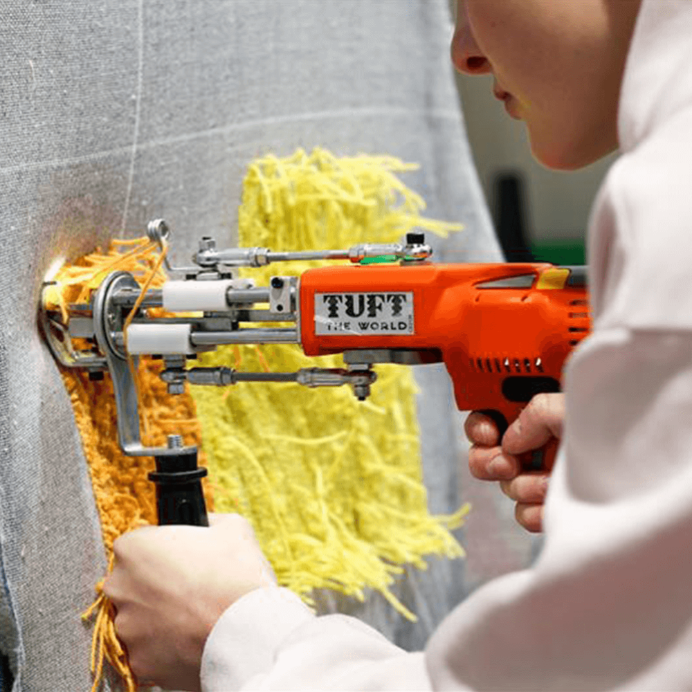 A student using an electric tufting gun to create a yellow and orange design on a piece of fabric.