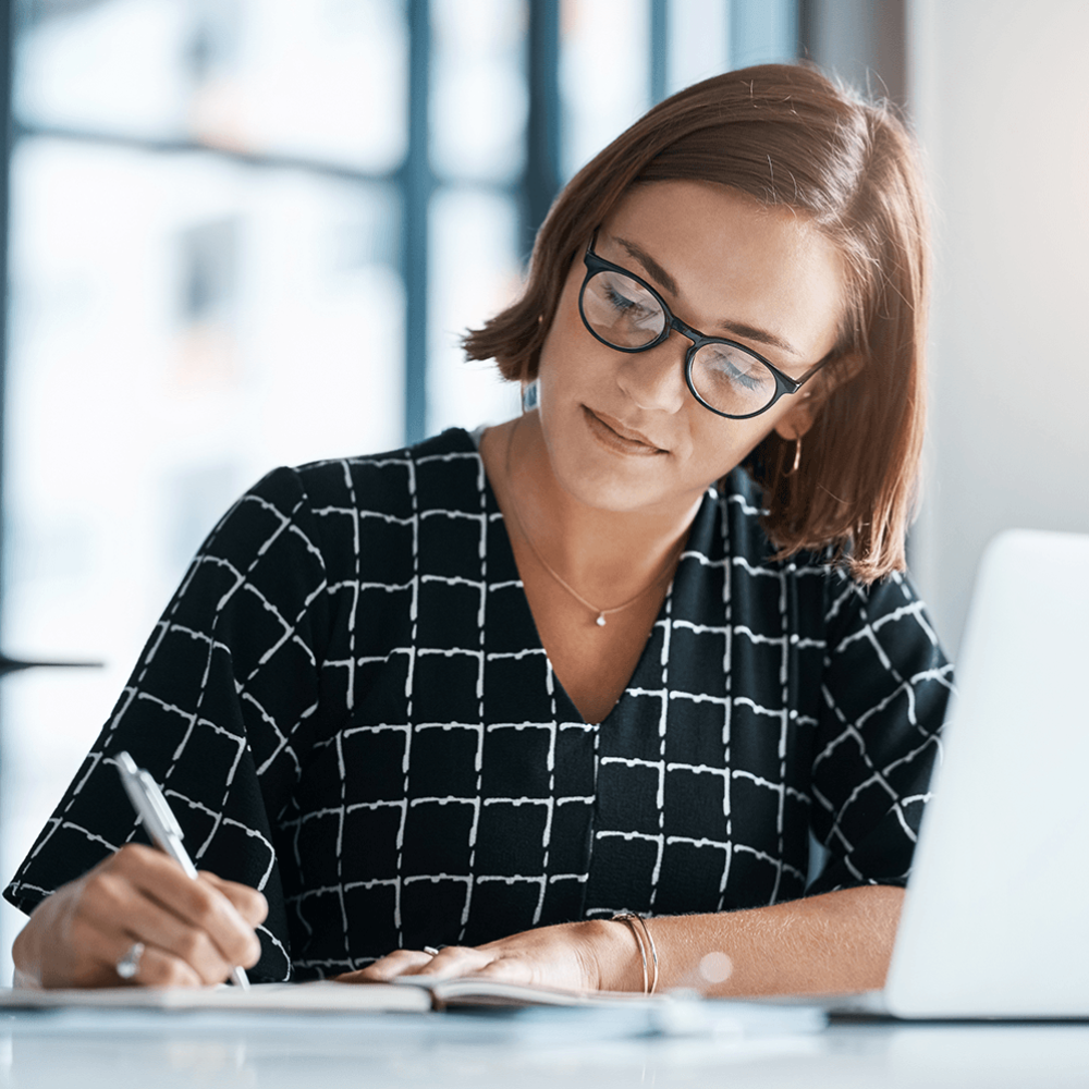A woman sat at her desk looking at her laptop and writing in a notebook