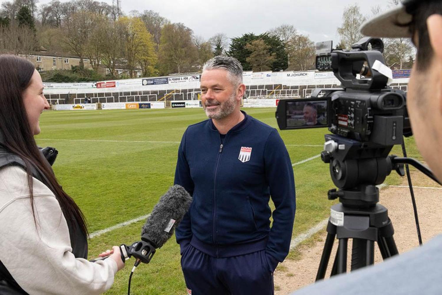 A student interviewing a sports personality on a football pitch as another student records it on camera