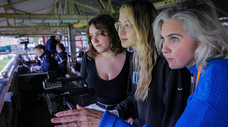 A group of women filming in a sports stadium.
