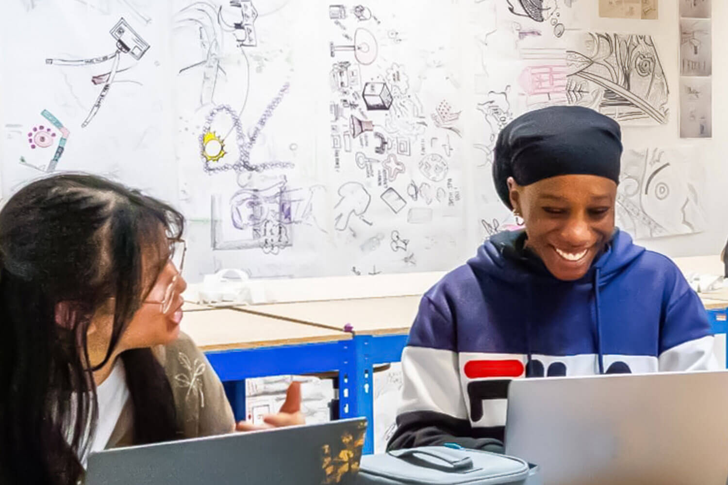 Two students chatting over laptops with illustrations pinned to the wall behind them