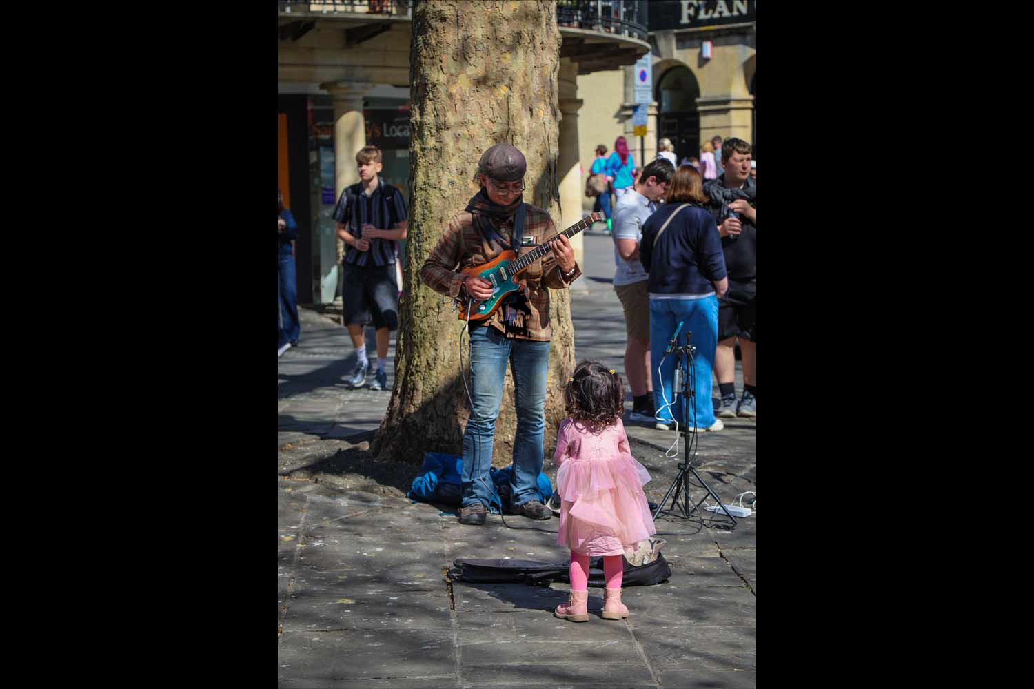 Winner of 2025 international photo comp. Little girl in pink dress gazes up at busker in central bath