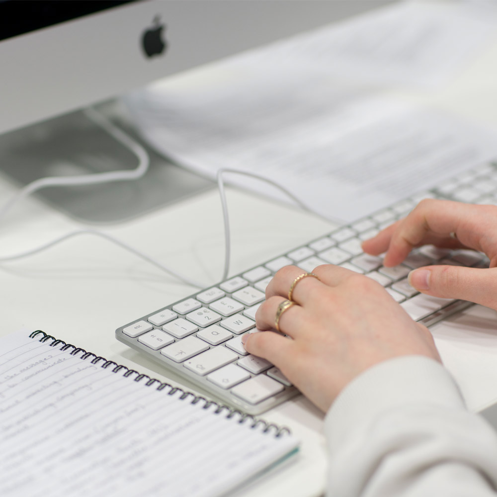 A closeup shot of hands typing on a desktop computer keyboard with a notepad of notes next to the keyboard on the desk.