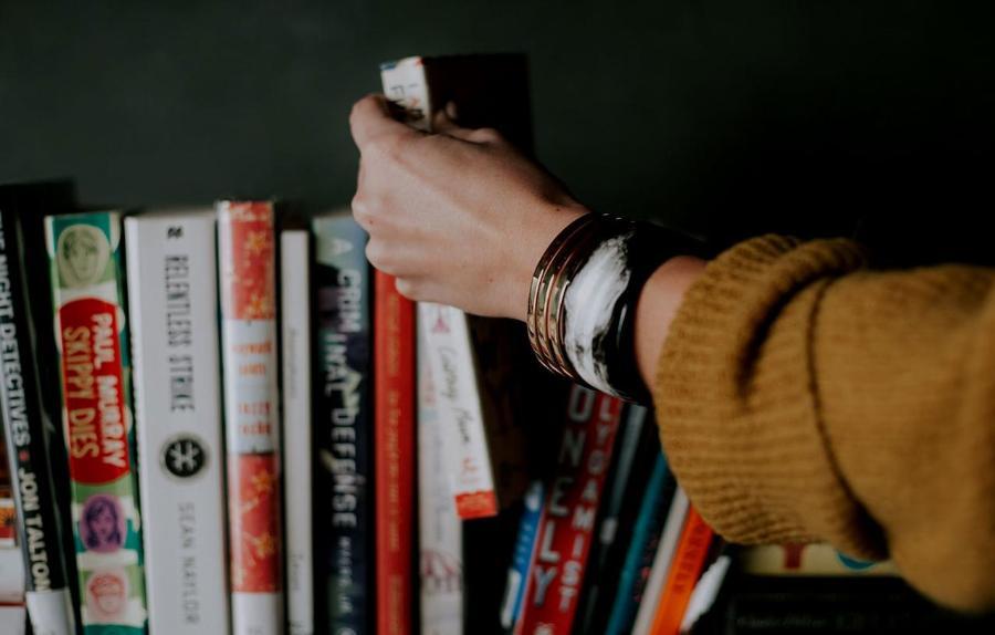 A woman's hand reaching towards a bookshelf to remove a book.