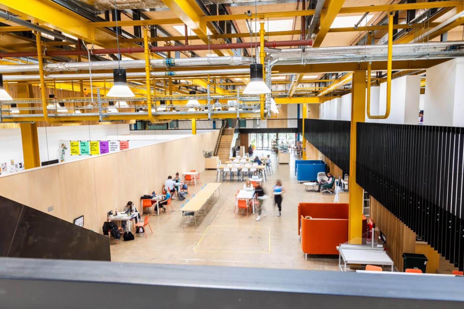An overhead shot of the main interior foyer of Locksbrook campus. The ceiling is covered in a range different coloured wires and pipes, and the foyer floor has an array of seats. Students walk through the centre of the image.