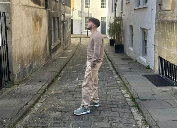 a man stood in a beige outfit on a cobbled street in front of bath stone buildings