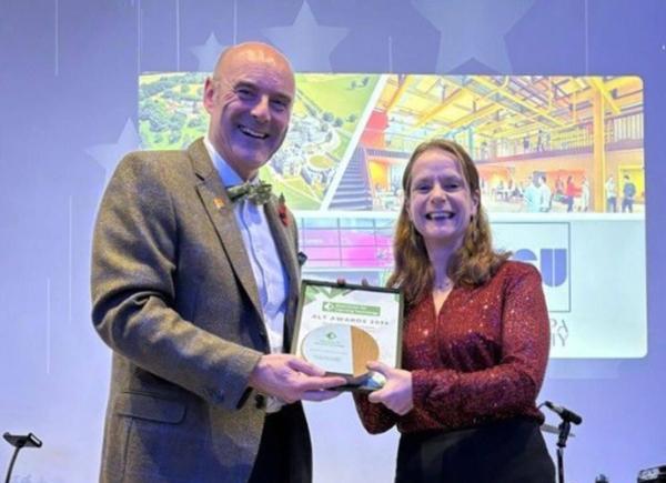 Two people stand on a stage, holding an award and certificate, in front of a BSU branded image on a backlit screen.
