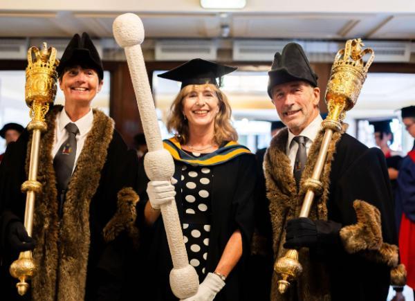 Arlene Stone dressed in academic robes, smiles as she holds the Ceremonial Mace and poses with the Mayor of Bath