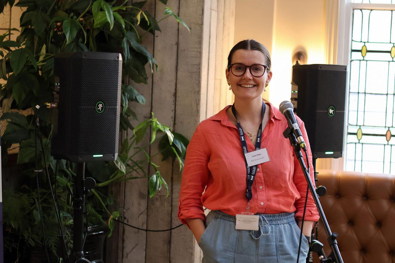 Lizzy stands in front of speakers and a plant at a BSIN event smiling to the camera.