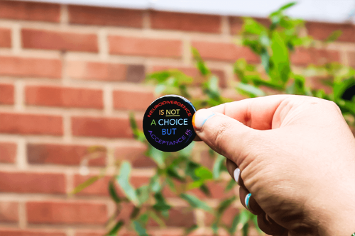 A hand holding up a black pin badge with text that says Neurodivergence is not a choice but acceptance is in rainbow letters.