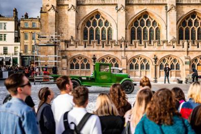 People gather to watch a green car driving by the Bath abbey