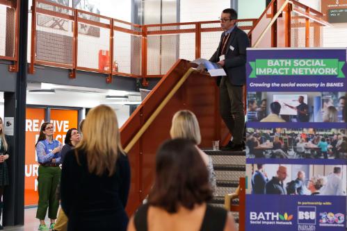 A person speaking at an event with a banner behind them