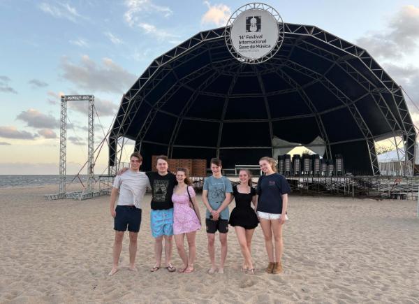 A group of students pose for a photo on a beach in front of a stage