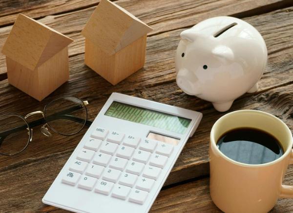 calculator, cup of coffee, piggy bank, wooden houses and glasses spread out on a wooden surface.