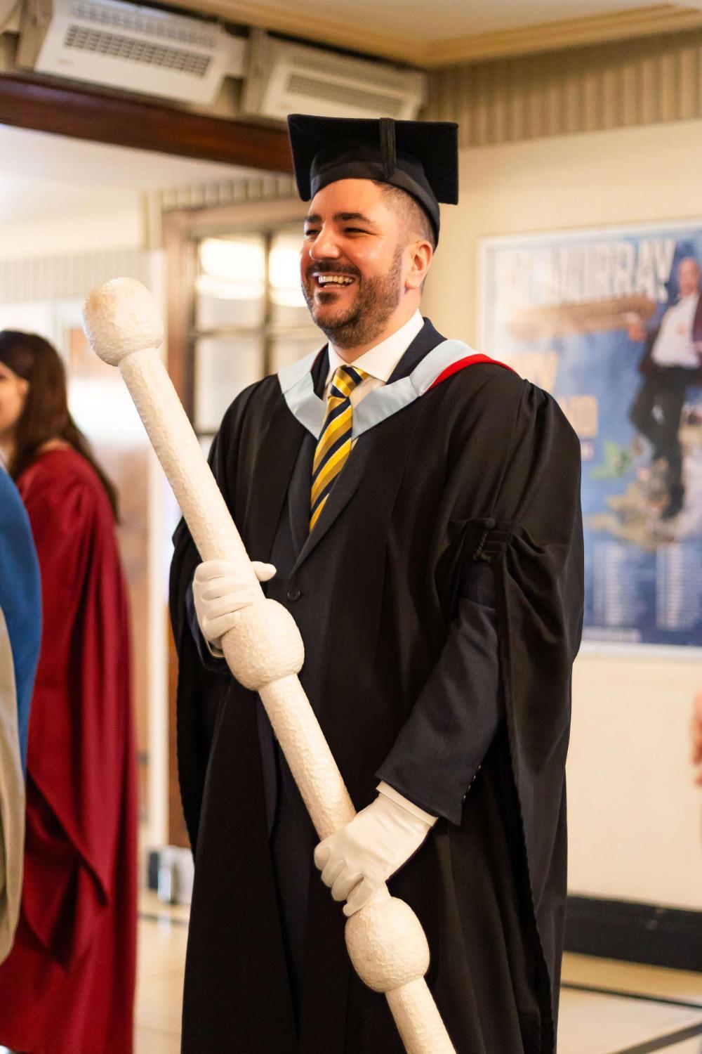 David Newman dressed in academic robes, smiling as he holds BSU's Ceremonial Mace