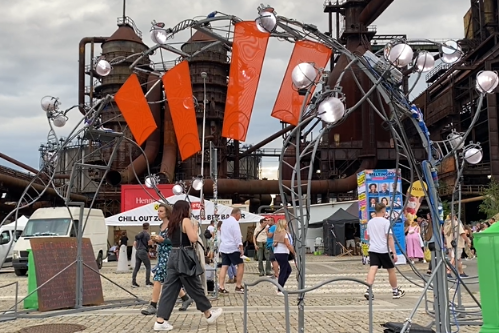 A crowd of people walking around at a festival, with a gate made out of random metal pieces and bright red flags on it.