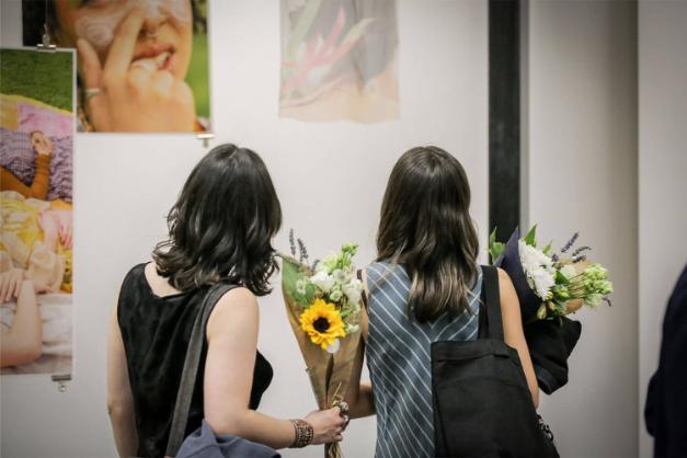 Two women with black hair admire a piece of their art with their back to the camera, both holding a bunch of flowers