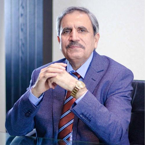 Man sits at a desk wearing a suit and a striped tie. His elbows are on the table and hands together.