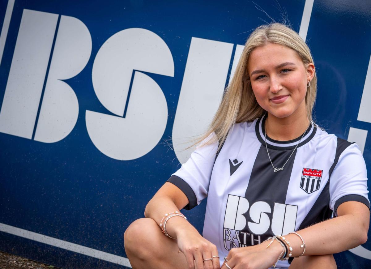 BSU Student Esme wearing a black and white Bath City shirt in front of a big BSU logo