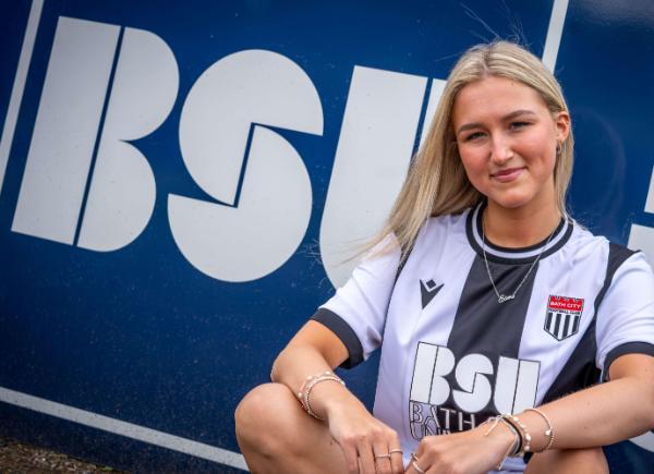 BSU Student Esme wearing a black and white Bath City shirt in front of a big BSU logo