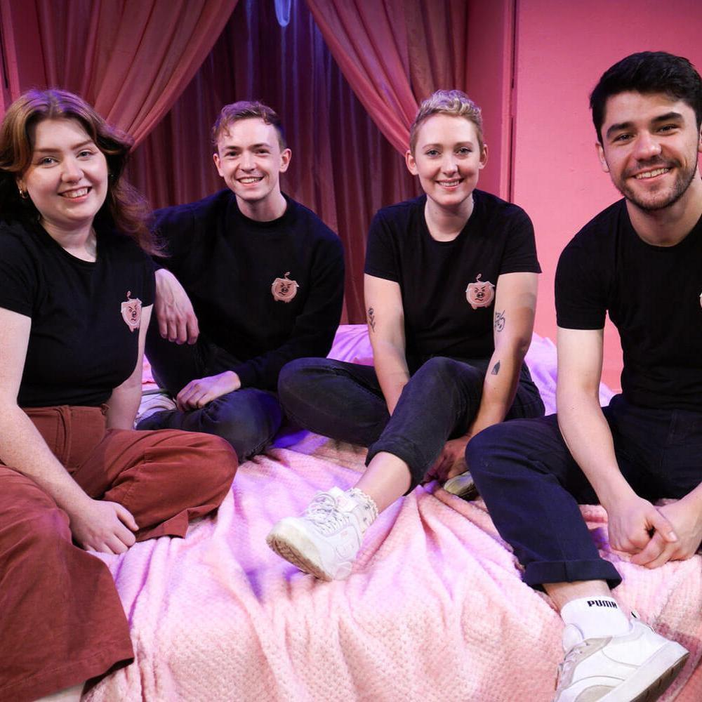 Four people sit on a bed, all dressed in black T-shirts, embroidered with cartoon pigs