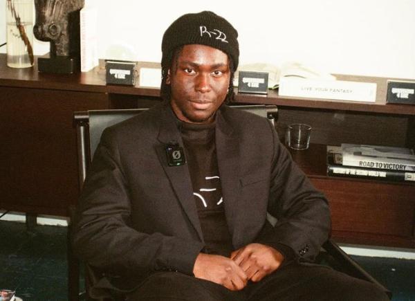 Hansel Abakah Phillips sitting in a chair, wearing a stylish suit and a beanie hat