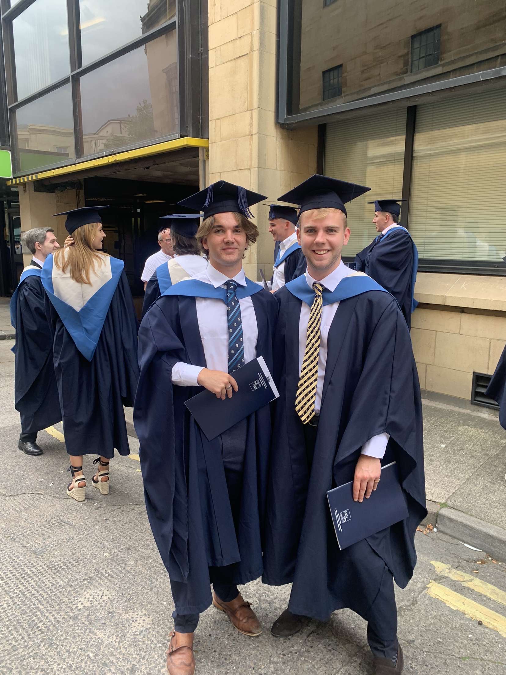 Two students in graduation cap and gowns outside a tall building