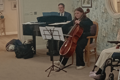 A female student dressed in black sits on a chair playing the bass violin. Behind her is a male student dressed in a black suit playing a grand piano.