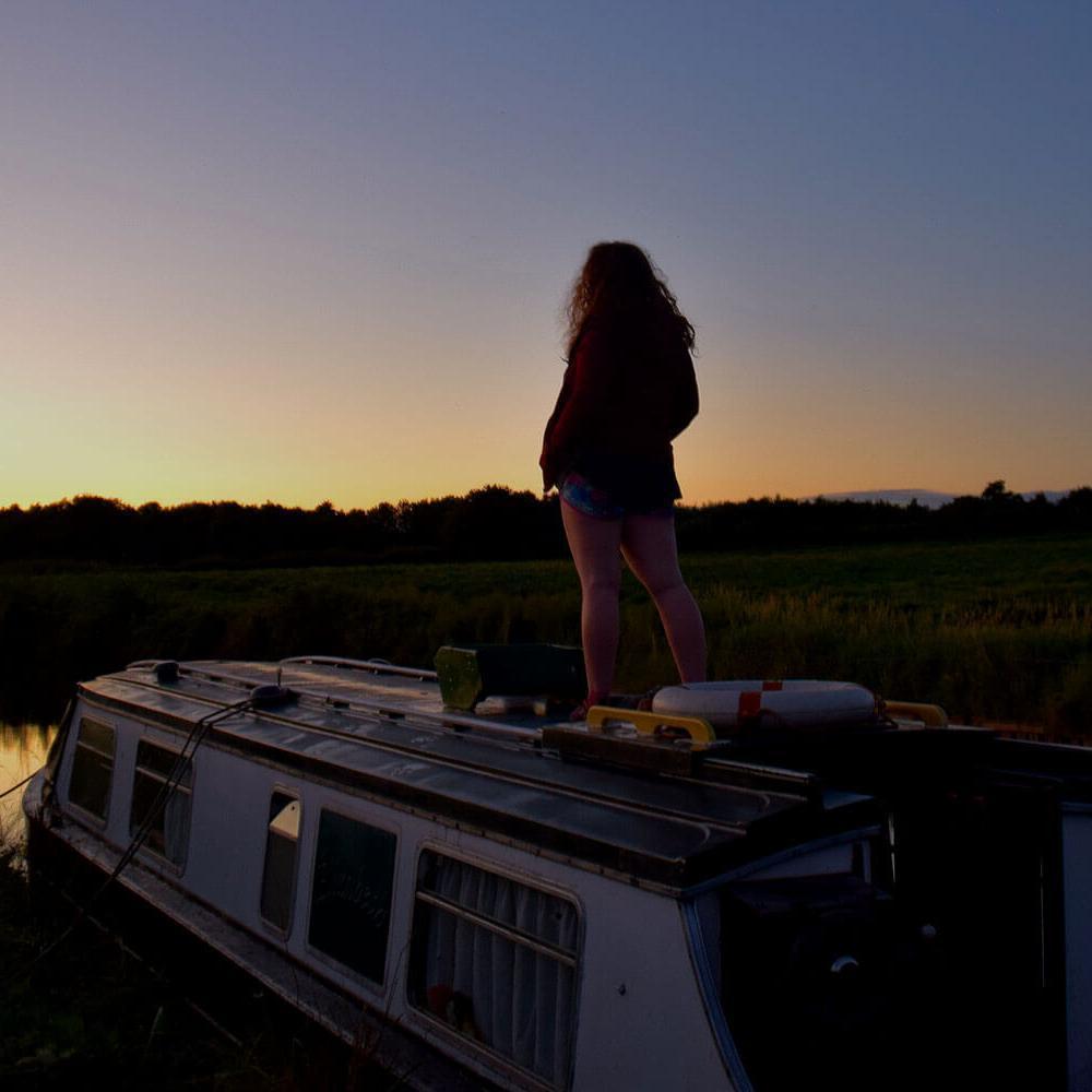 A woman stands on top of a narrowboat as it sails down a canal at sunset.