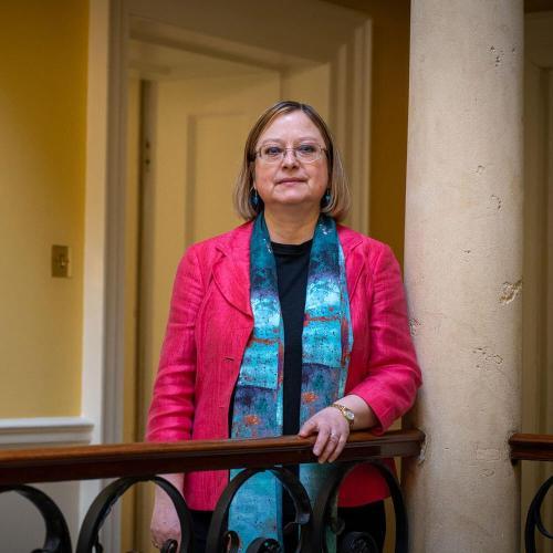 A woman in a pink jacket stands in a hallway with one hand on a banister