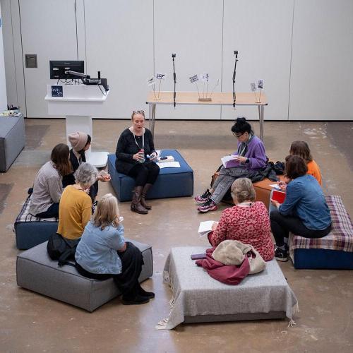 people sitting in a circle in the centre of a white room on cushions listening to one of the people talking.