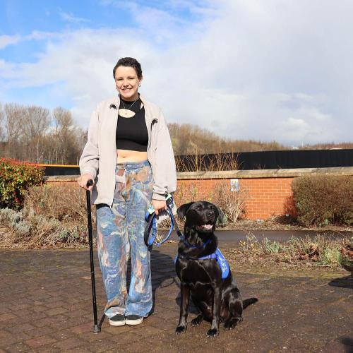 A woman with short black hair and a walking sticks hold a black support dog on a lead and smiles